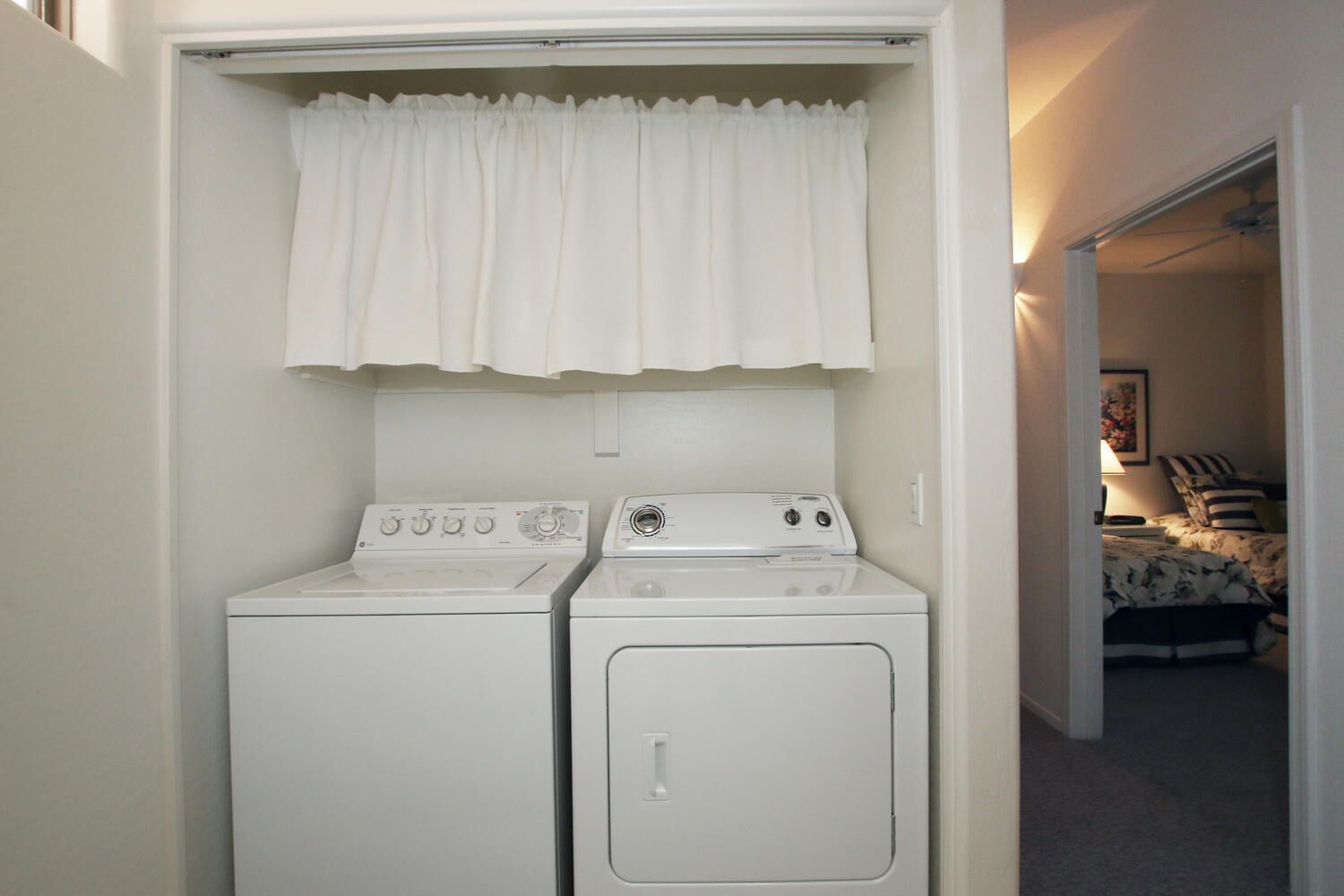 A laundry room with two white washers and dryers, a white curtain, and a bed visible through an open door.