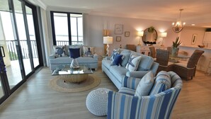 Amplia sala de estar con un sofá grande, una mesa de centro y un sillón a rayas. Cuenta con un comedor con mobiliario de mimbre y un candelabro.