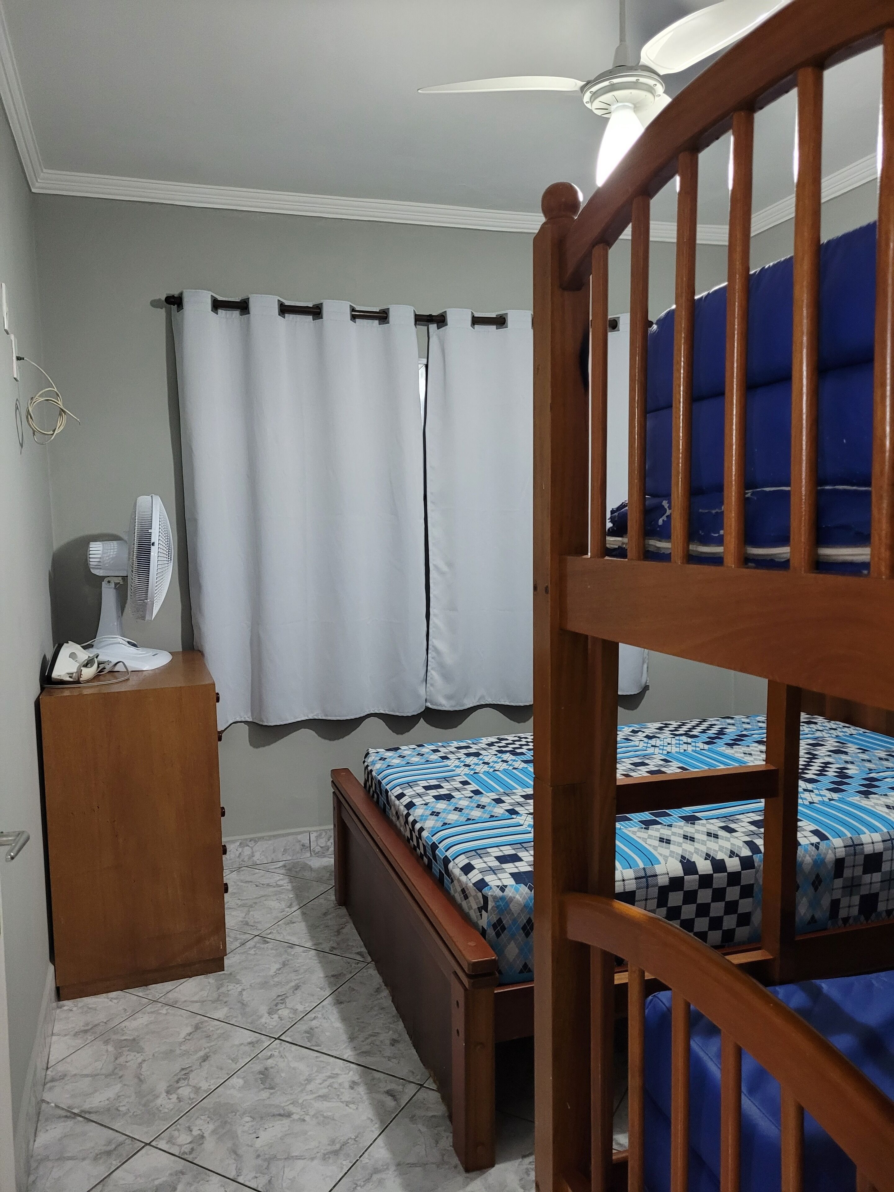 A bedroom with a wooden bunk bed, a single bed with a patterned bedspread, a wooden nightstand with a fan, a curtain, and a ceiling fan.