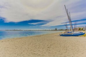 A sandy beach with a calm sea, a sailboat, and a distant cityscape.