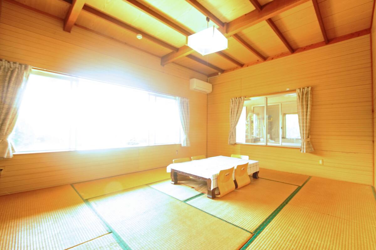 Ein Zimmer mit Tatami-Boden, einem Bett, einem Vorhangfenster und einem Deckenventilator.