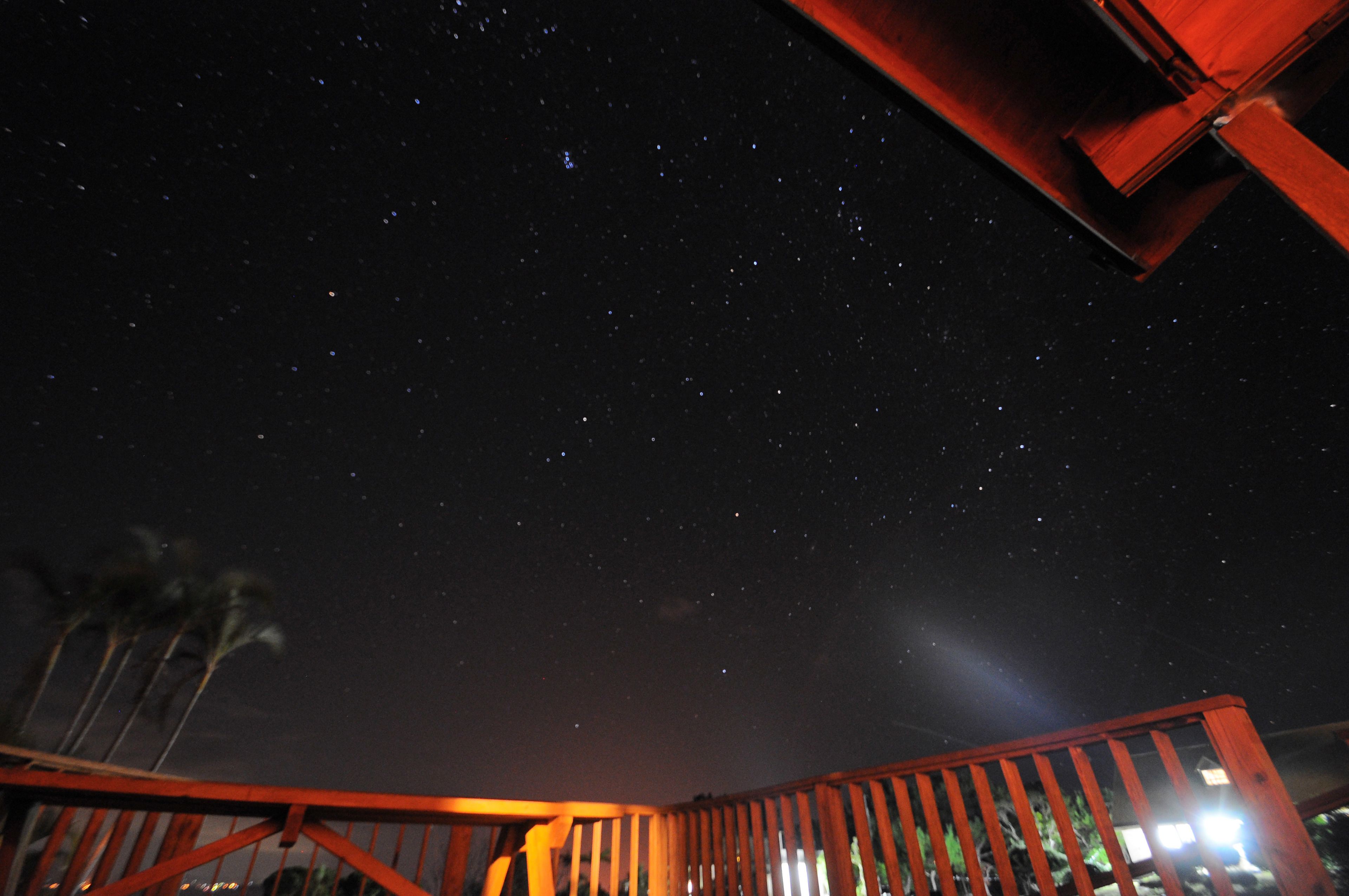 Cielo nocturno lleno de estrellas, visto desde una terraza de madera con barandal rojo.