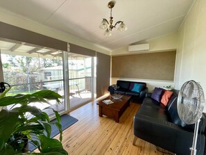 Een ruime woonkamer met een schuifdeur naar een terras, een zwarte leren bank, een houten salontafel en een staande ventilator.