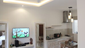 A modern kitchen with a built-in oven, a dining area with a glass table, and a TV displaying a cartoon. - Green Studio Seminyak (Kuta)