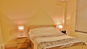 A hotel room with a single bed, two bedside lamps, a nightstand, and a window with blinds. - Beautiful 2 Bedroom Private Apartment (London)