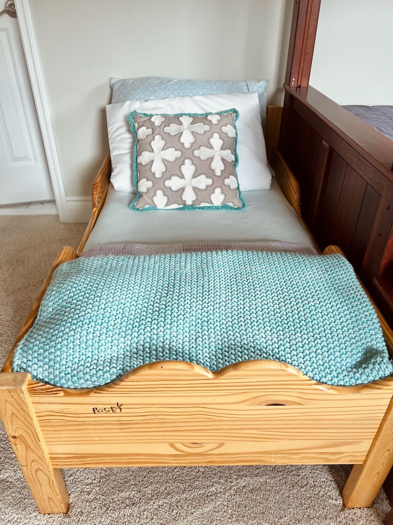 A wooden bed with a teal knitted blanket, a pillow with a floral pattern, and a white pillow.