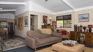 A spacious living room with a beige sofa, a coffee table, and a dining area with chairs. There is a large window with a view of the outdoors.
