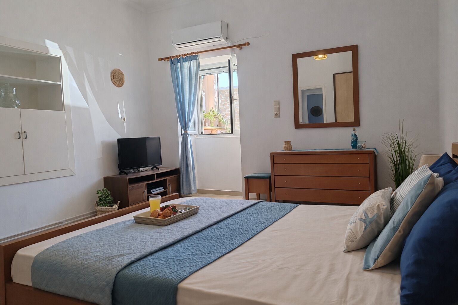Un dormitorio con cama, tocador, televisor, ventana con cortinas y espejo en la pared.