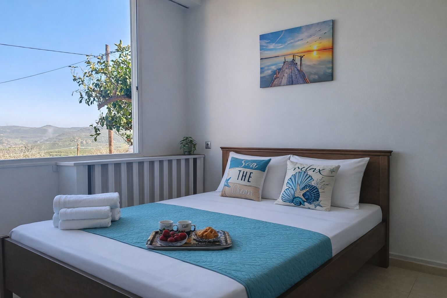 Ein Schlafzimmer mit Bett, Kissen, einer Ablage mit Snacks, einem Gemälde und Blick ins Freie.