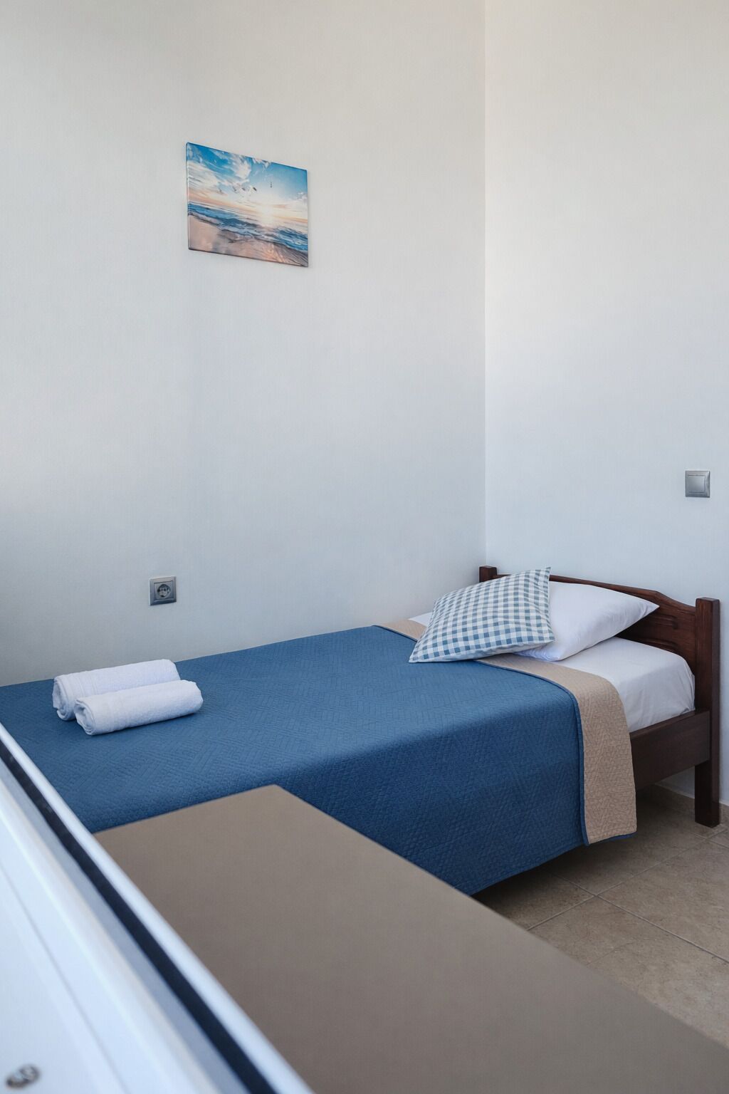 Een enkel bed met een blauwe spreidekker en een geruit kussen, een klein nachtkastje en een wandcontactdoos.