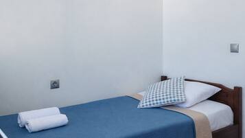 A single bed with a blue bedspread and a checkered pillow, a small bedside table, and a wall-mounted power outlet.
