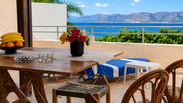 Ein Essbereich mit Tisch, Stühlen, einer Obstschale, einer Blumenvase und Blick auf das Meer.