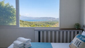 Ein Schlafzimmer mit einem großen Fenster, das einen Blick auf eine Küstenlandschaft bietet, einem ordentlich bezogenen Bett mit blauer Steudecke und einem Kissen mit einem Seestern-Motiv.