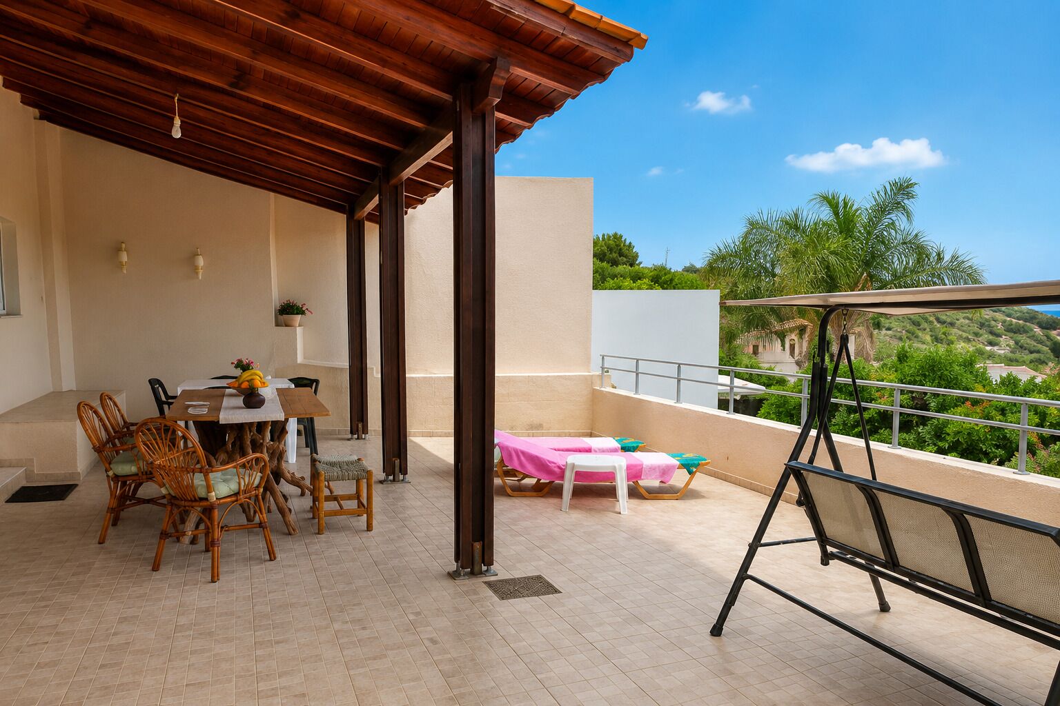 Eine überdachte Terrasse im Freien mit Tisch und Stühlen, ein kleiner Tisch mit einer Vase sowie Blick auf Grünflächen und Hügel.