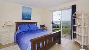 A bedroom with a bed, a nightstand, a painting, a window with a view, and a white shelf unit.