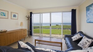 A living room with a view of the sea, a wooden cabinet, a table, and a sofa with patterned cushions.