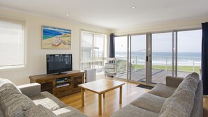 A spacious living room with a television, a wooden coffee table, and a sofa. There is a large window with a view of the ocean.