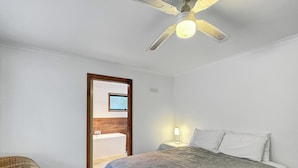 Une chambre à coucher avec un lit, une table de chevet, une lampe, un ventilateur de plafond, une chaise en osier et une salle de bain visible à travers une porte ouverte.