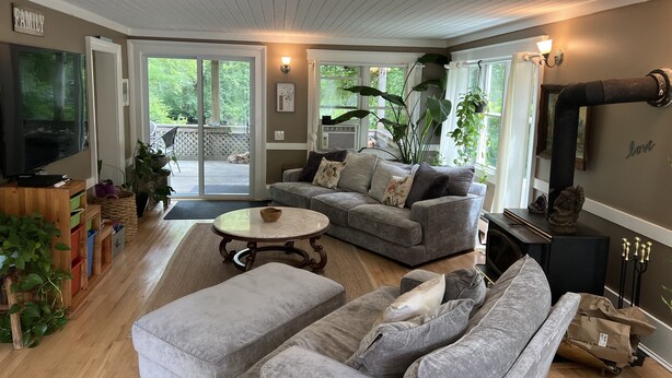 A spacious living room with a grey sofa, a coffee table, and a television. There are large windows and a view of the outdoors.