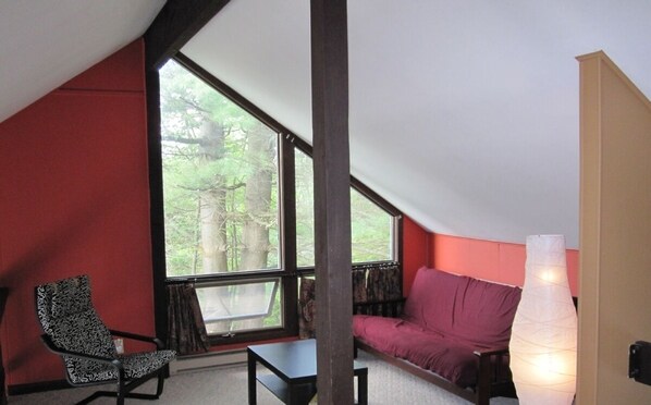 Una habitación con techo inclinado, un ventanal grande, pared roja, una silla estampada, una mesita pequeña, un sofá morado y una lámpara alta y blanca.