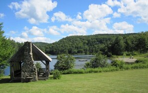 A stone gazebo with a thatched roof overlooking a lake, surrounded by lush greenery and a forested hill in the background. - Spacious/Elegant 3 level house on Spruce Lake- swim or fish, short drive to Mt. (Wilmington)