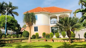 A two-story house with a red-tiled roof, a balcony on the upper floor, and a well-maintained garden with palm trees and shrubs. - Beautiful two-level, fully furnished 4 bedroom house with WiFi (Kampala)