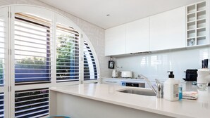 A modern kitchen with white cabinetry, a built-in dishwasher, and a large window with blinds. - Balmoral Beauty - DIRECT BEACHFRONT - Romantic Studio Escape (Mosman)