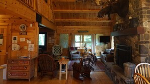 A rustic cabin interior with a stone fireplace, wooden walls, a mounted moose head, and a cozy seating area.