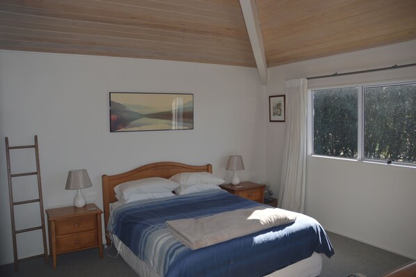 A bedroom with a wooden ceiling, a bed with a blue and white bedspread, two bedside tables with lamps, a ladder, a window with a view of trees, and a painting on the wall.