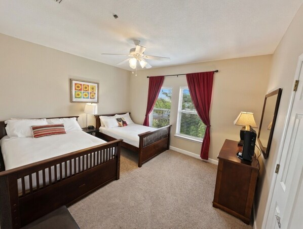 A bedroom with two beds, a ceiling fan, a window with curtains, a bedside table with a lamp, and a framed picture on the wall.