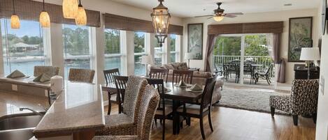 A spacious living area with a dining table, wicker chairs, a kitchen island, and large windows offering views of the outdoors.