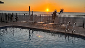 Une piscine avec vue sur le coucher de soleil sur l’océan, une plage avec des chaises longues et une clôture.