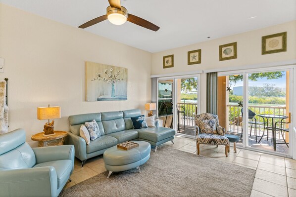 A spacious living room with a light blue sofa, a patterned armchair, and a coffee table. There is a ceiling fan, a painting on the wall, and a view of the outdoors through a sliding glass door.
