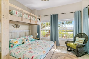 Quarto com uma cama beliche de madeira, uma cama de solteiro com roupa de cama colorida, uma cadeira de vime e vista para o verde através de uma janela.