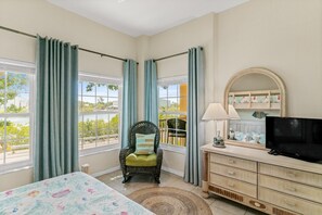 Une chambre à coucher comprenant un lit, une chaise, une commode, un téléviseur et de grandes fenêtres avec des rideaux.