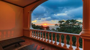 A balcony with a view of the sunset over the sea, a wooden bench, and a balustrade.