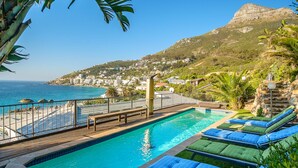 A swimming pool with lounge chairs, a wooden deck, and a view of the sea and a hill. - Breathtaking Clifton 4th beach villa (Clifton)
