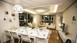 Un coin repas avec une table blanche dressée pour six personnes, entourée de chaises blanches. On aperçoit au fond un canapé blanc, une grande fenêtre donnant sur la ville et un plafond orné d’un motif décoratif.