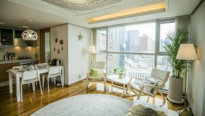 Un salon moderne avec un coin repas, de grandes fenêtres offrant une vue sur la ville, un plafond orné de motifs complexes et une cuisine en arrière-plan.