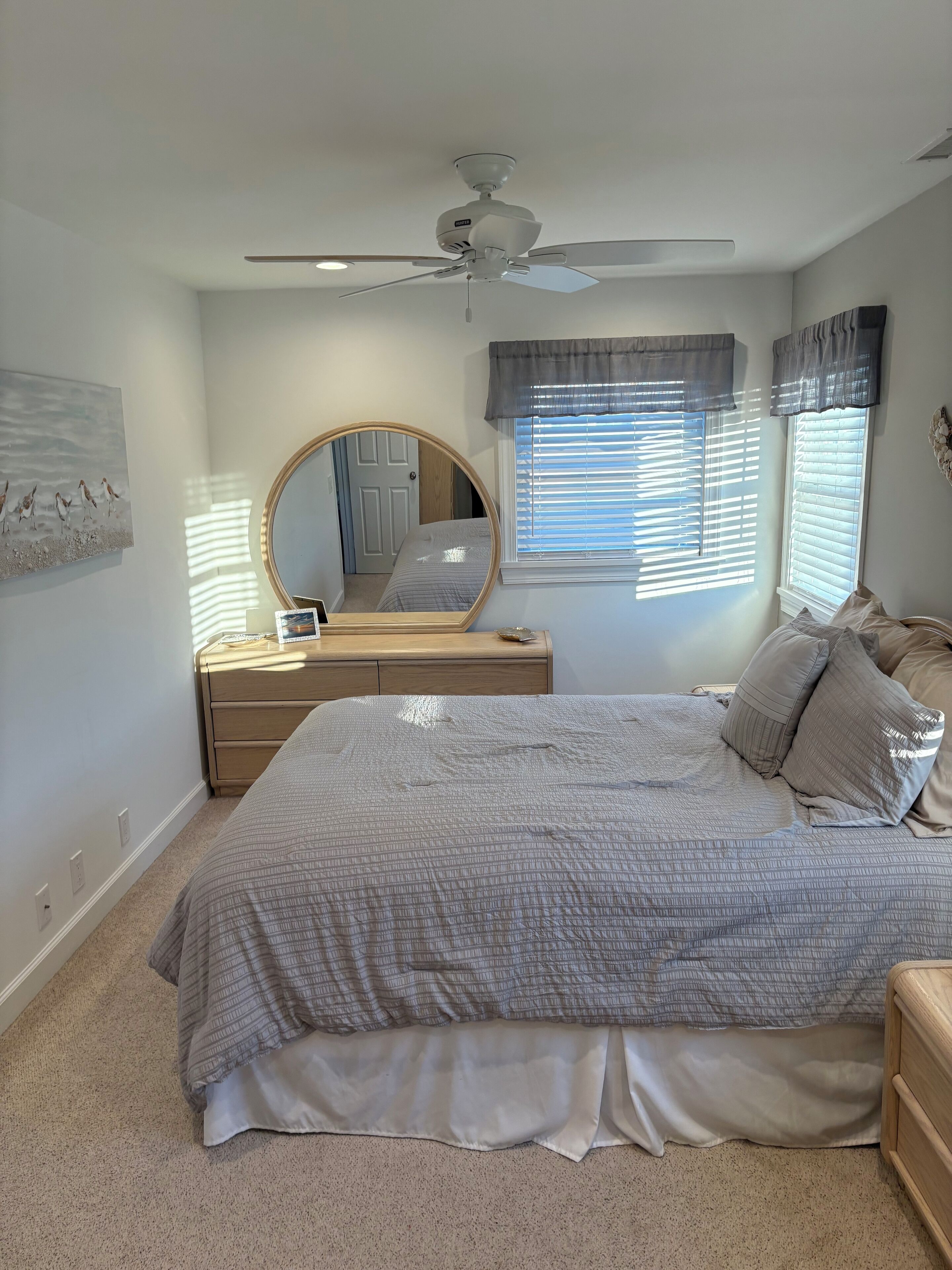 Une chambre à coucher comprenant un lit, un miroir rond, une table de chevet, un ventilateur de plafond et des fenêtres équipées de stores.
