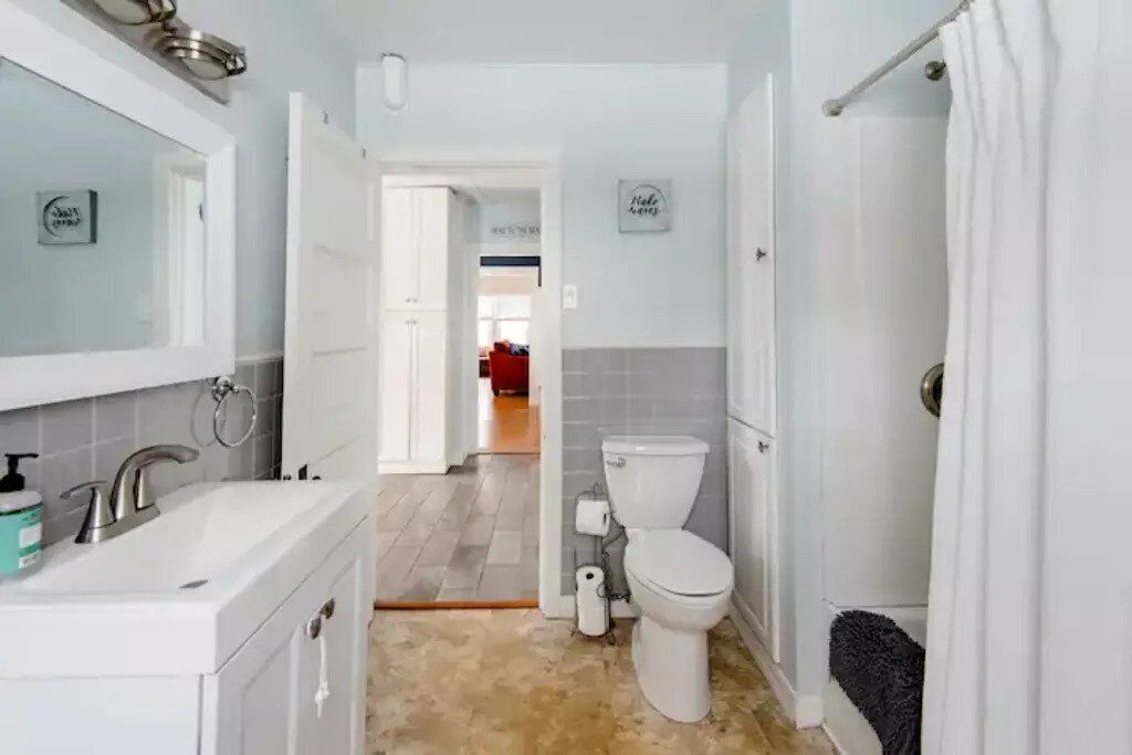 Un baño con lavamanos blanco, espejo y retrete. Hay un perchero y una cortina de ducha.