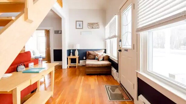 A cozy living room with a wooden staircase, a sofa, a small table, and a window with blinds. - Adorable Beach House Across the Street From Beach! (Scarborough)