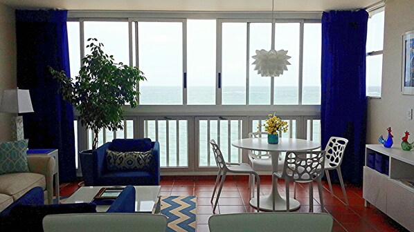 Une pièce avec vue sur la mer, une table à manger avec des chaises, un canapé, une plante en pot et une petite table ornée d’objets décoratifs.