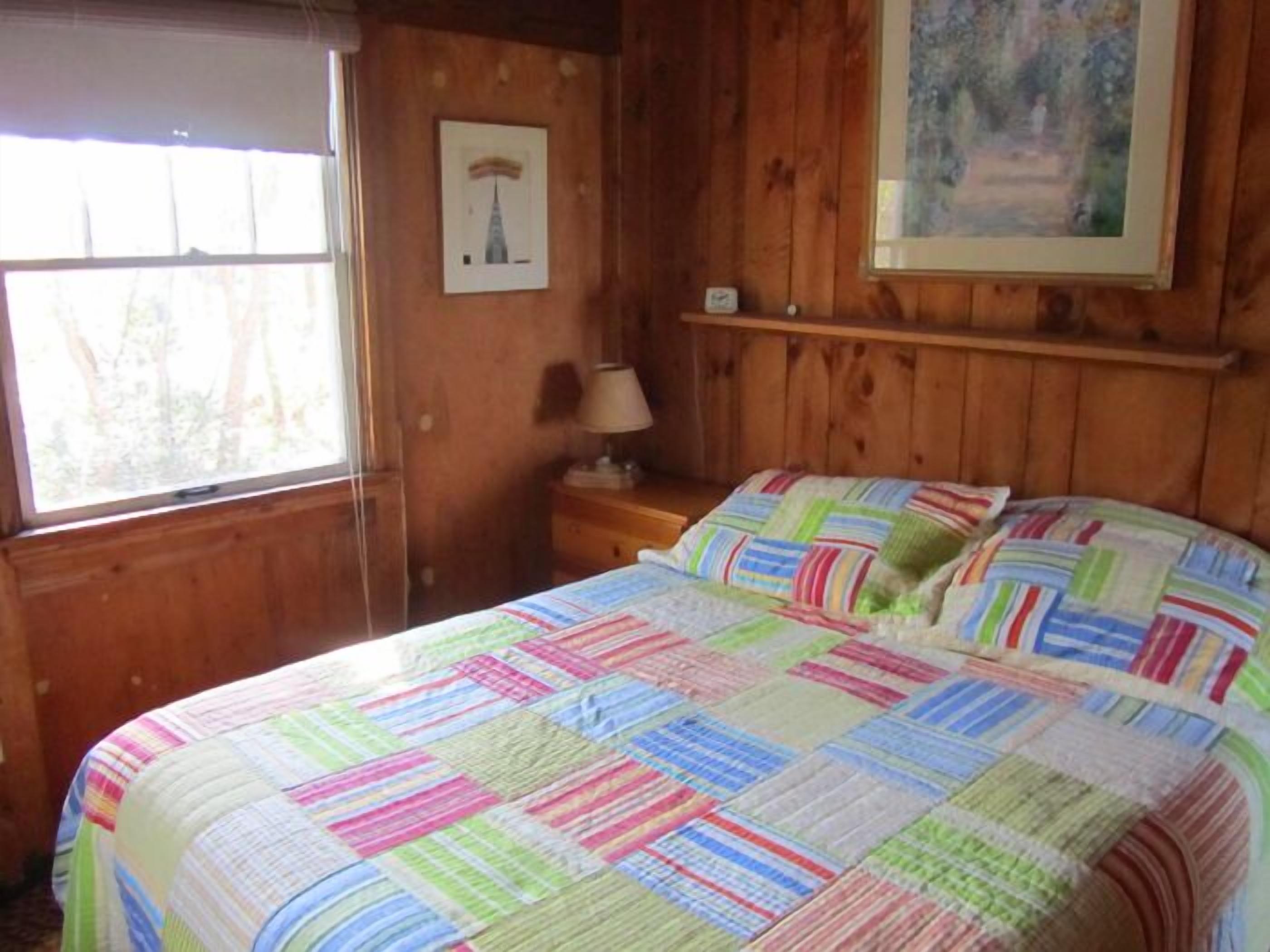 Une pièce de style cabane en bois, avec un lit, une table de chevet, une lampe, un tableau encadré au mur et une fenêtre.