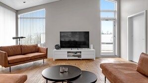 Een moderne woonkamer met een grote flatscreen-tv op een witte tv-meubel, twee bruine leren banken, een ronde salontafel en grote ramen met jaloezieën.