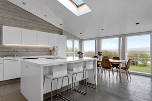 A modern kitchen with a central island, a dining area with a table and chairs, and large windows offering a view of the outdoors. - Luxury Villa with river view In South Iceland - The Golden Circle (Laugarás)