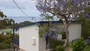 A small, single-story house with a balcony, surrounded by greenery and a tree with purple flowers.