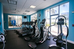 A gym with treadmills, ellipticals, and a stationary bike, large windows, and a clock on the wall.
