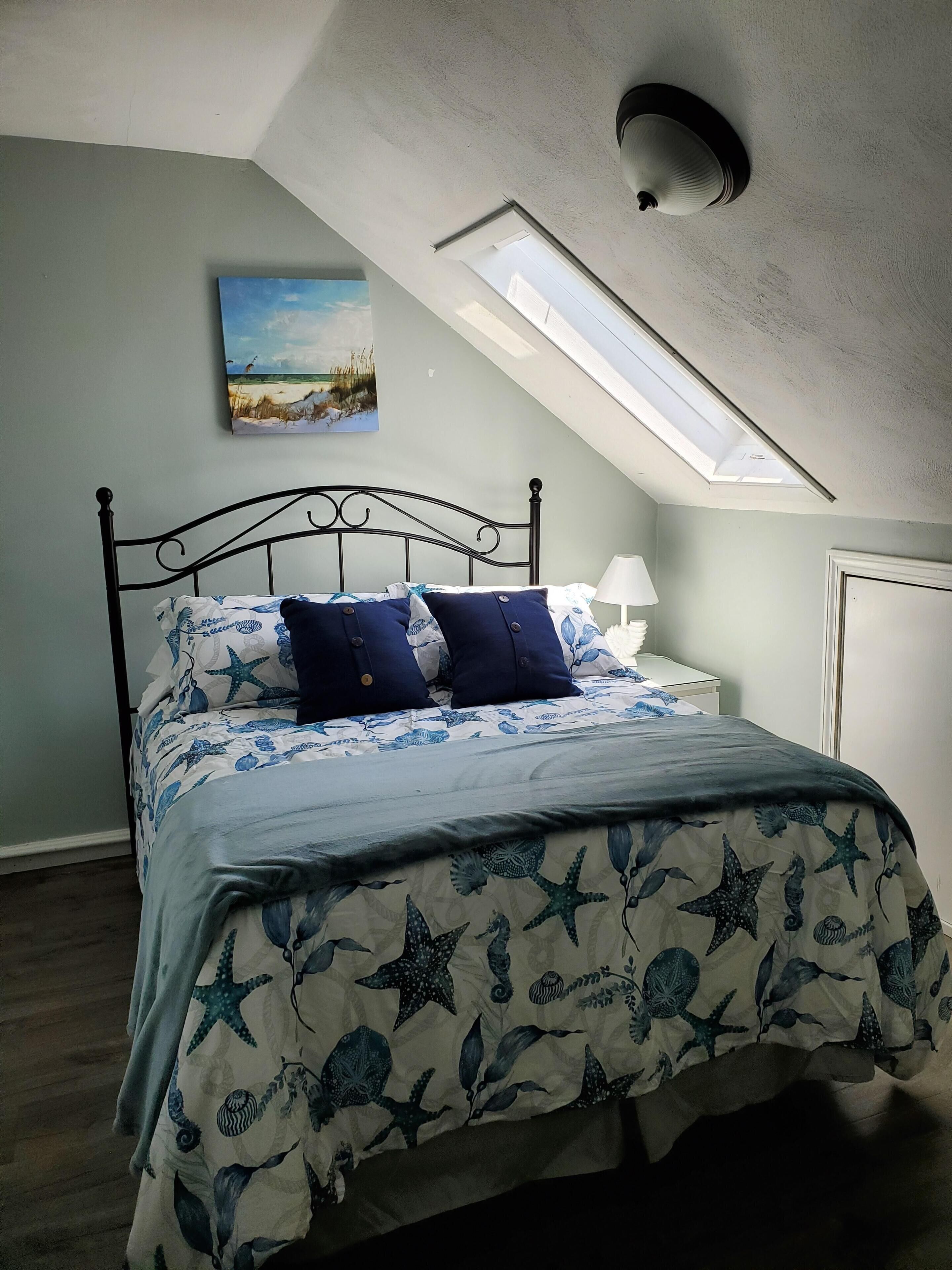 A bedroom with a sloped ceiling, a skylight, a bed with a starfish and seahorse themed bedspread, a nightstand with a lamp, and a painting of a beach scene.