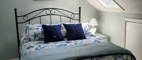 A bedroom with a sloped ceiling, a skylight, a bed with a starfish and seahorse themed bedspread, a nightstand with a lamp, and a painting of a beach scene.